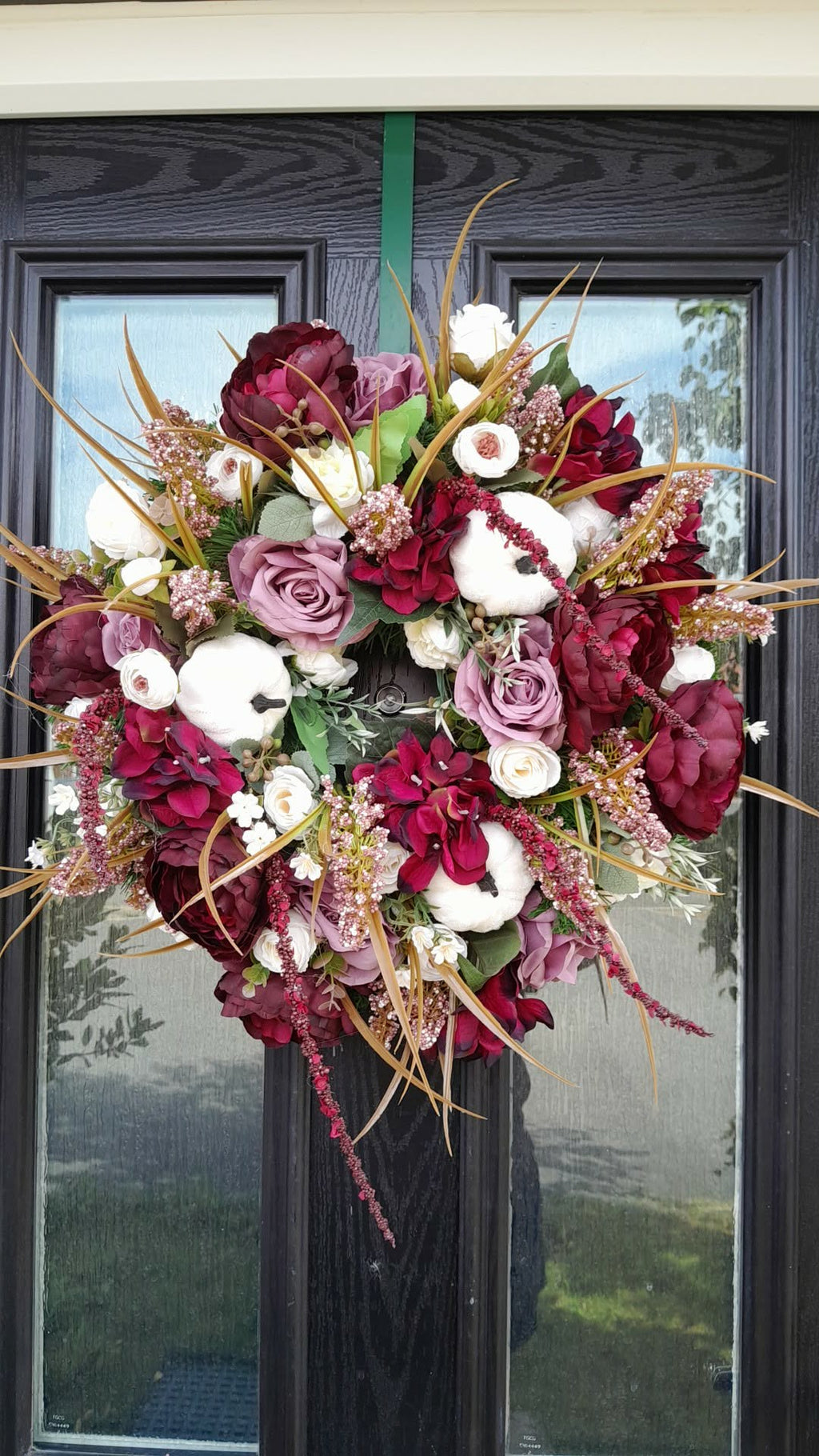 Autumn Burgundy and Mauve Luxury Wreath hanging on a black front door, featuring deep burgundy peonies, mauve roses, white ranunculus, cotton stems and golden dried grasses