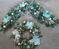 Teal and silver Christmas 6ft garland and matching wreath set laid out on a grey floor with teal magnolia blooms, silver baubles and snowflake details